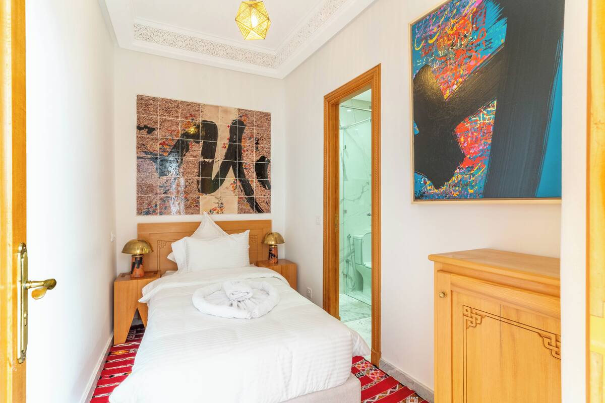 A hotel room with a bed, a wooden nightstand, a red patterned rug, a wooden cabinet, and two paintings on the wall.