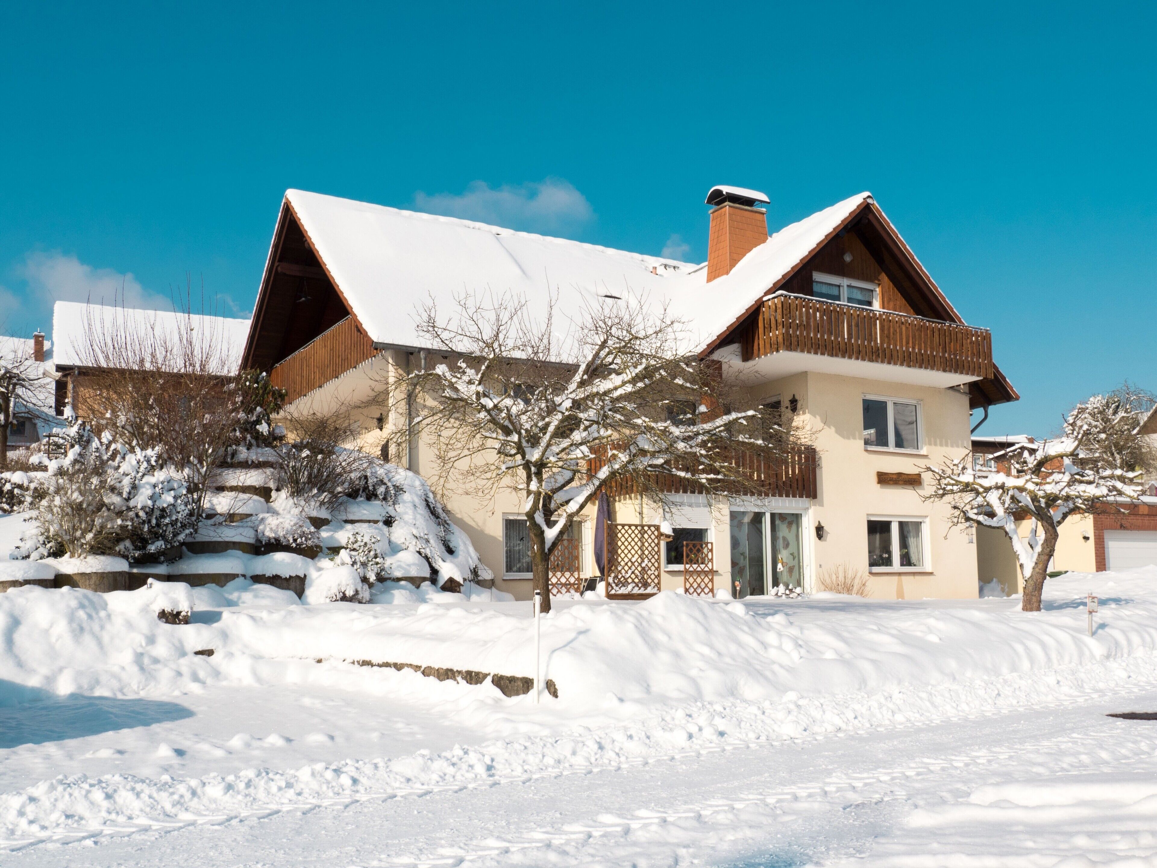 A two-story house with a snow-covered roof, a wooden balcony, and a stone staircase leading to the entrance.