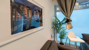 Ein Zimmer mit großem Fenster, Blick auf eine Stadtlandschaft, einer Pflanze, einem Schreibtisch mit Stuhl und einem Sofa.
