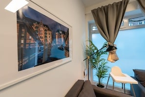 A room with a large window, a view of a cityscape, a plant, a desk with a chair, and a sofa. - Apartment 2 Hamburg Winterhude (Hamburg)