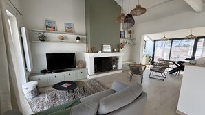 Een moderne woonkamer met een grote flatscreen televisie, een open haard en een eethoek met rieten stoelen.
