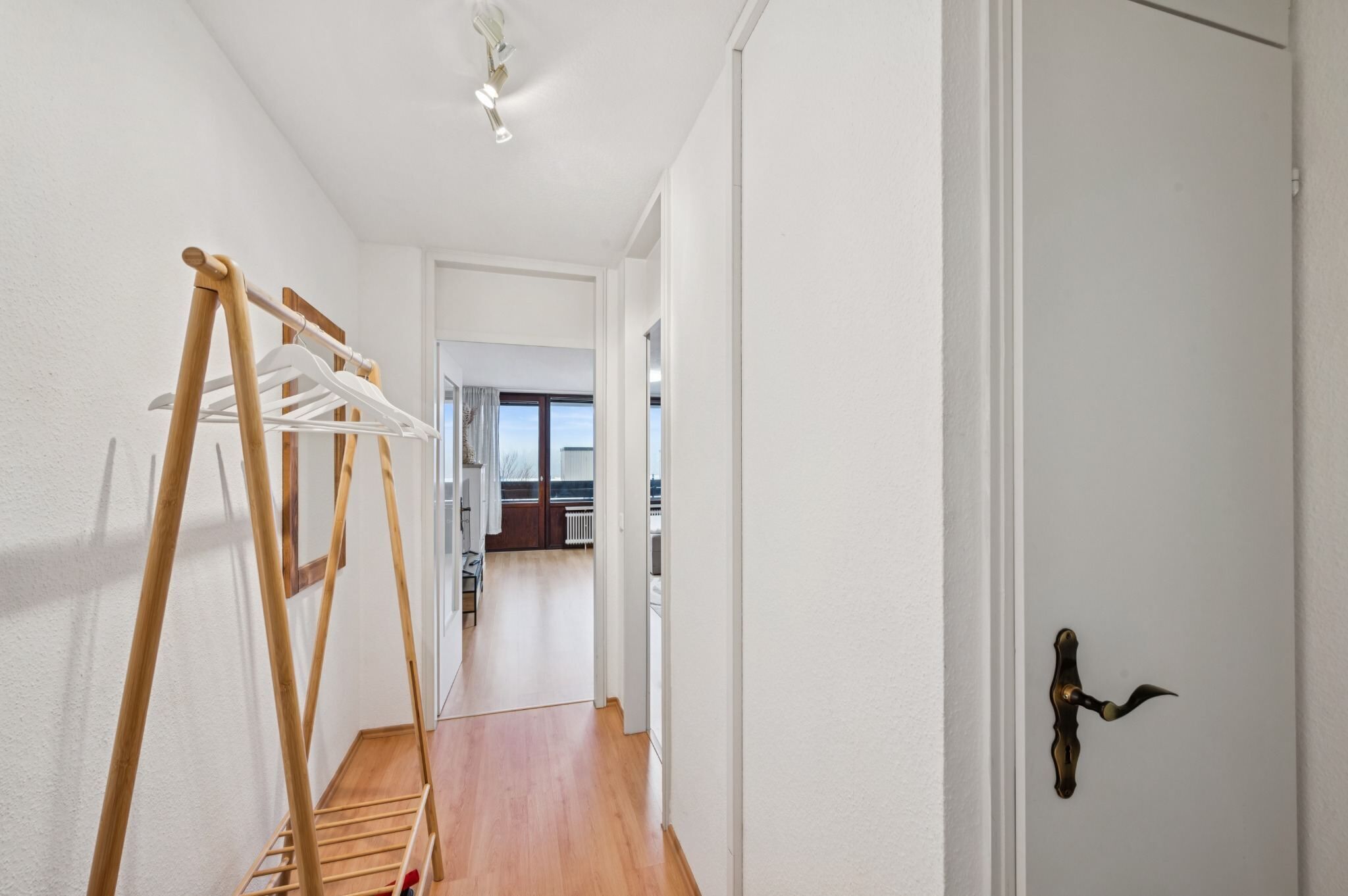 Un couloir avec un étendoir en bois, une porte blanche avec une poignée en laiton, et une vue sur une pièce avec une fenêtre.