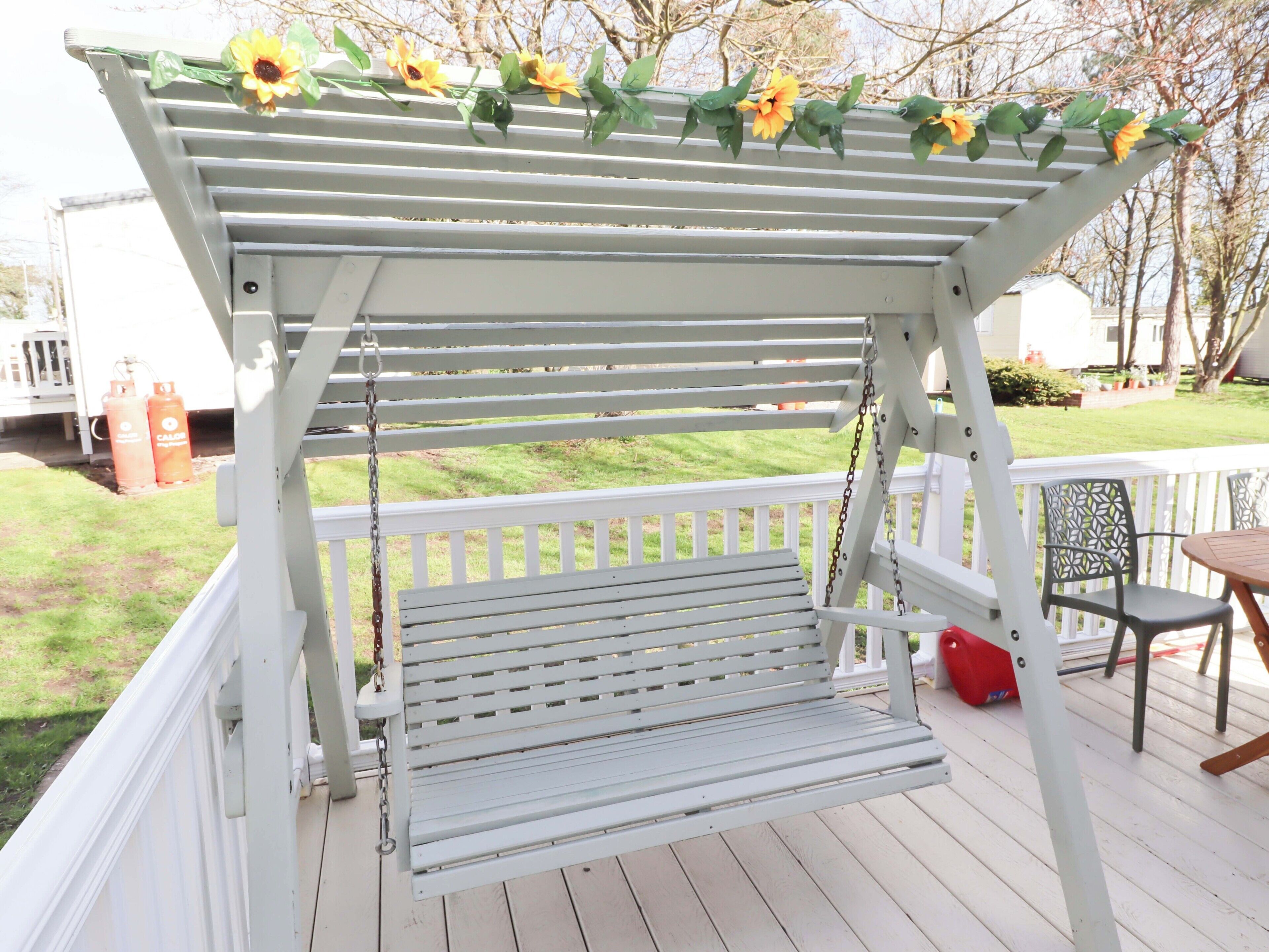 Une balançoire extérieure blanche, avec un banc en dessous, entourée d’une balustrade blanche et décorée de tournesols.