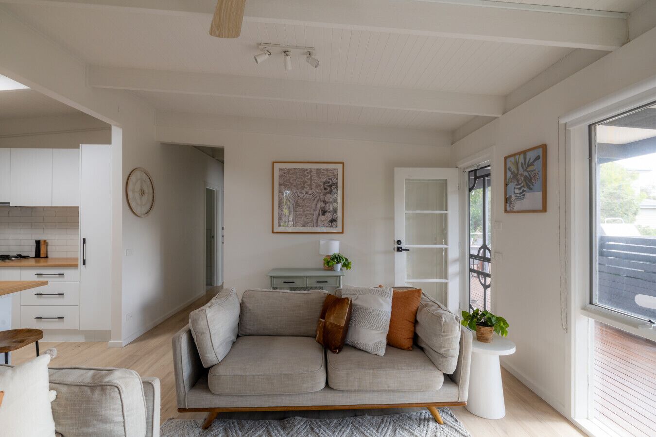 A modern living room with a sofa, a coffee table, and a side table with a plant. There is a kitchen area with cabinets and a countertop visible.