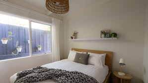 A bedroom with a bed, bedside table, lamp, window with curtains, and a shelf with plants. - Lovely Beach Cottage Heart of Rye (Rye)