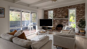 A living room with a brick fireplace, a television, and a large window overlooking a deck. - Lovely Beach Cottage Heart of Rye (Rye)