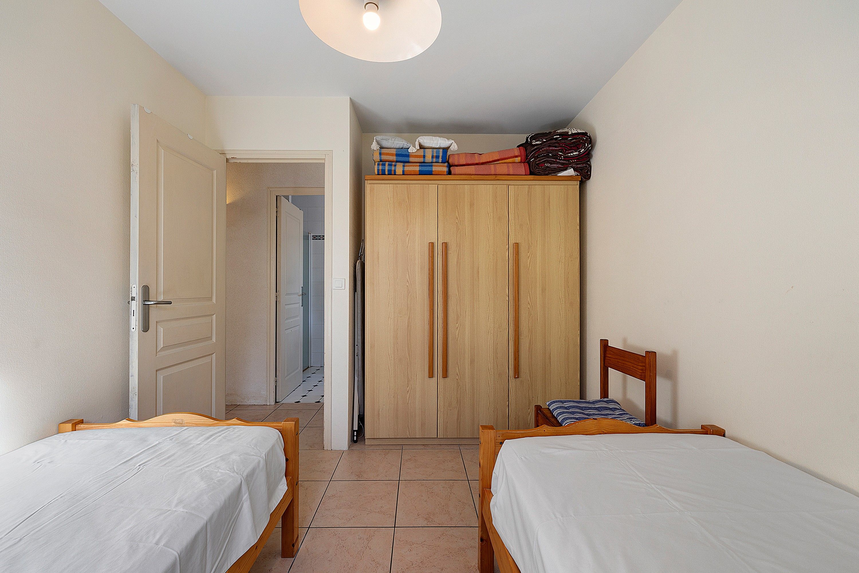 A small, well-lit room with two single beds, a wooden wardrobe, and a door leading to a bathroom.