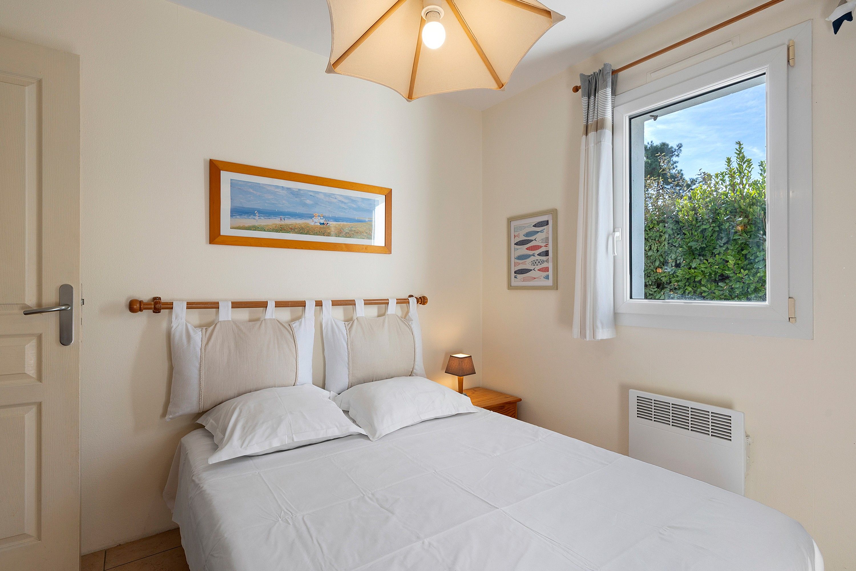 Un dormitorio con cama, ventana con vista a la vegetación, un cuadro en la pared y una lámpara de noche.