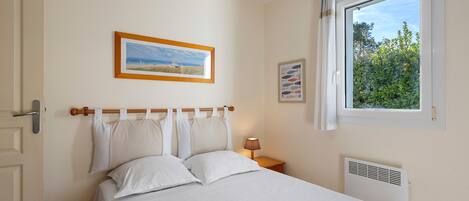 Un dormitorio con cama, ventana con vista a la vegetación, un cuadro en la pared y una lámpara de noche.
