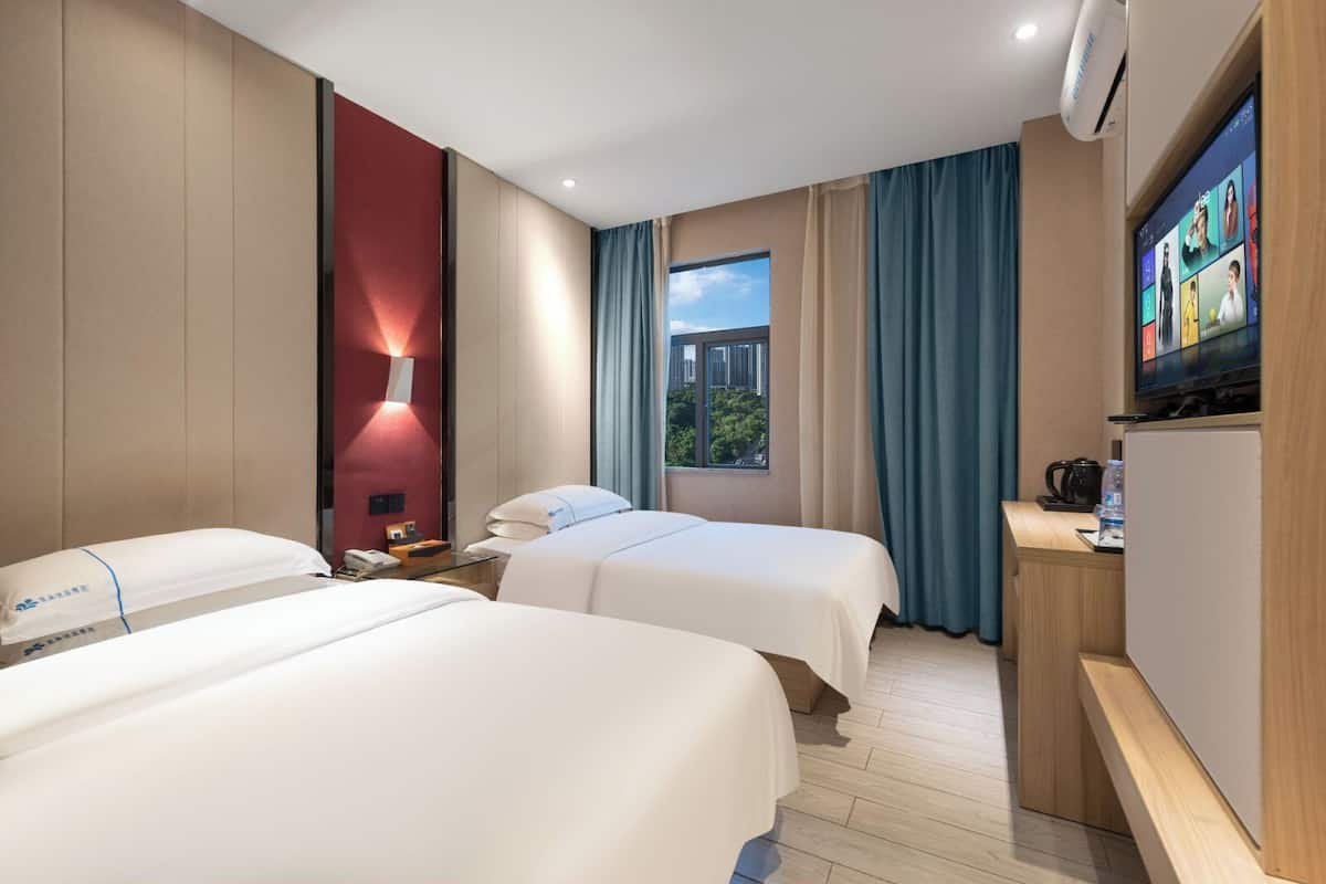 Quarto de hotel com duas camas, televisão, secretária e janela com cortinas, oferecendo vista para a cidade.