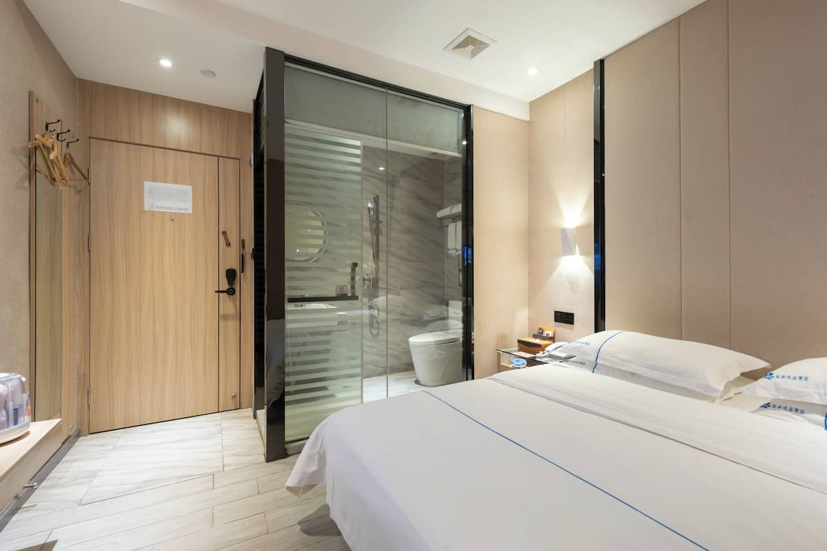 Quarto de hotel moderno com cama, guarda-roupa, casa de banho com cabine de chuveiro de vidro e mesa de cabeceira com candeeiro.