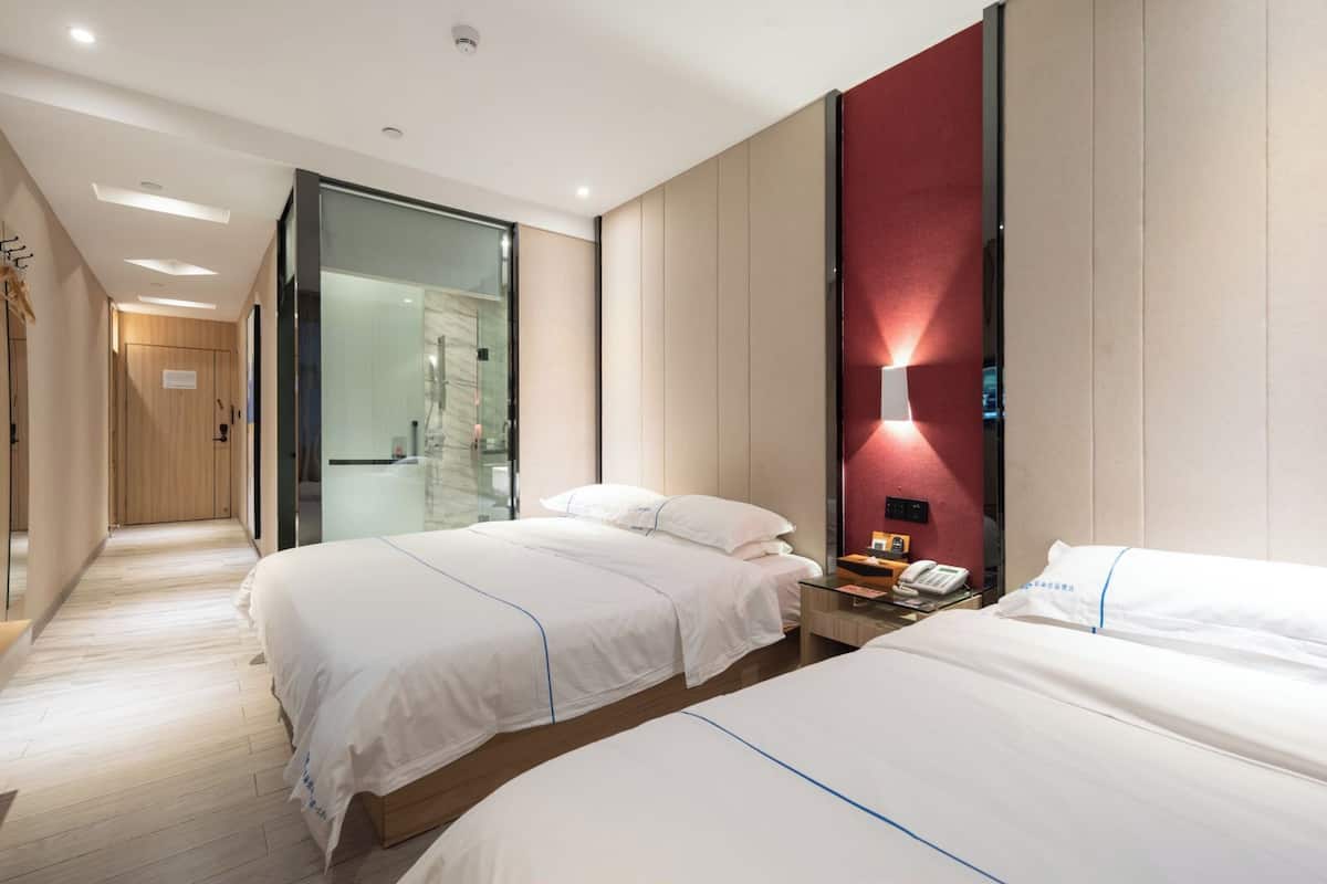 Quarto de hotel com duas camas, uma porta de vidro para um banheiro e uma parede com detalhes vermelhos.