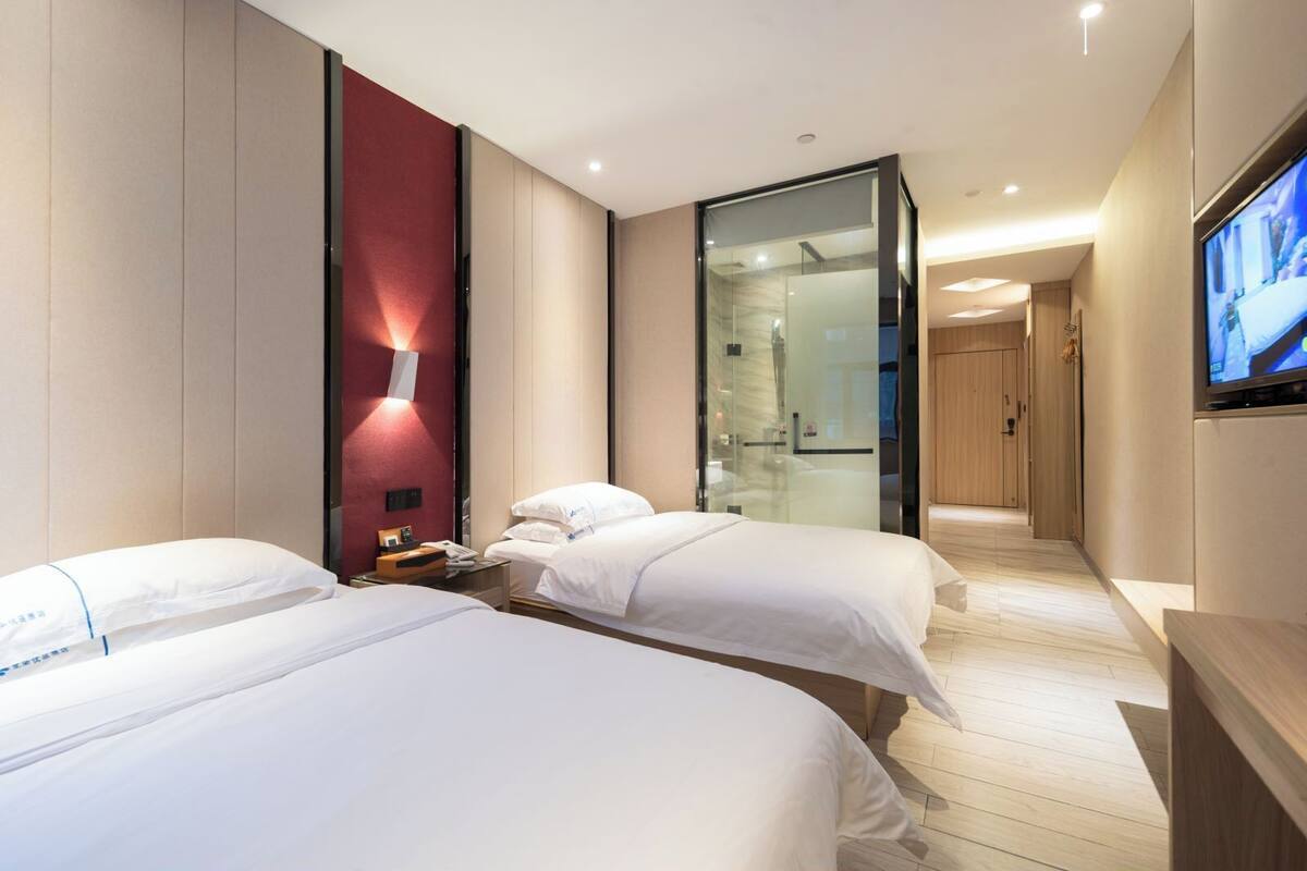 Quarto de hotel com duas camas, televisão, divisória de vidro e chão de madeira.