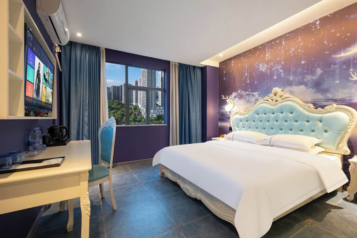 Quarto de hotel com cama, escrivaninha, cadeira, televisão e uma grande janela vista para a cidade.