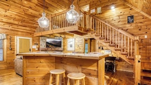A wooden cabin interior with a central bar area, wooden stairs, and a dining table. - Nestled Inn | Gated Forest Retreat | Hot Tub + 2 Jacuzzi Baths | Fireplace | (Sevierville)