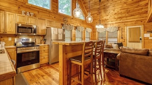A cabin interior with wooden walls, a kitchen with a microwave, stove, and refrigerator, a dining area with a table and chairs, and a sofa. - Nestled Inn | Gated Forest Retreat | Hot Tub + 2 Jacuzzi Baths | Fireplace | (Sevierville)
