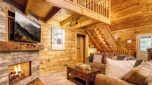 A log cabin interior with a stone fireplace, a wooden staircase, a sofa, and a television mounted on the wall. - Nestled Inn | Gated Forest Retreat | Hot Tub + 2 Jacuzzi Baths | Fireplace | (Sevierville)