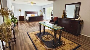 A spacious living room with a wooden floor, a glass coffee table, a ceiling fan, a bed with a headboard, and a large mirror. - Spacious Southside Home 1 King & 1 Queen bed! (Corpus Christi)
