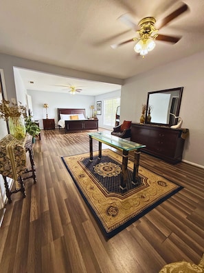 A spacious living room with a wooden floor, a glass coffee table, a ceiling fan, a bed with a headboard, and a large mirror. - Spacious Southside Home 1 King & 1 Queen bed! (Corpus Christi)
