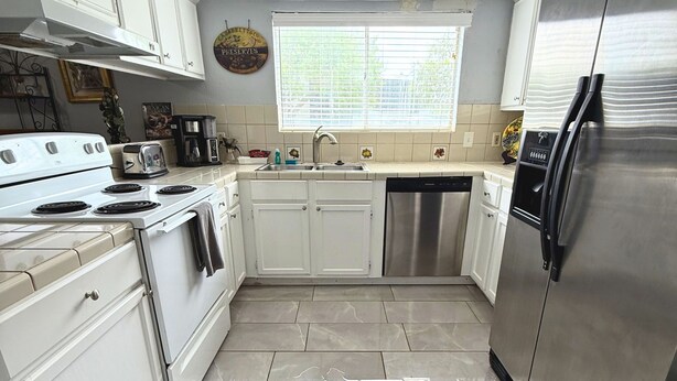 A modern kitchen with white cabinets, a stainless steel refrigerator, a dishwasher, and a window with blinds. - Spacious Southside Home 1 King & 1 Queen bed! (Corpus Christi)