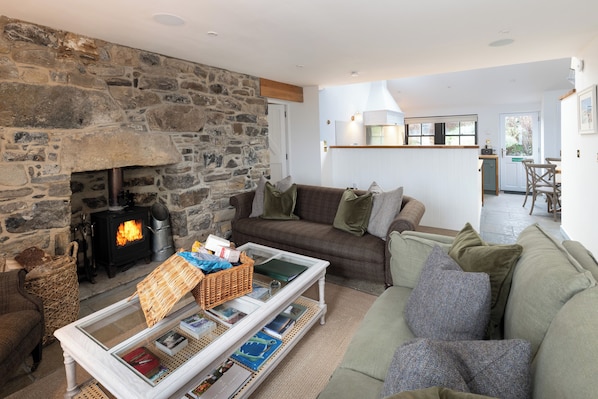 A cozy living room with a stone fireplace, a sofa, a coffee table with magazines, and a kitchen in the background. - Storytellers at Isay | Luxe Skye Coastal Stay (Waternish)