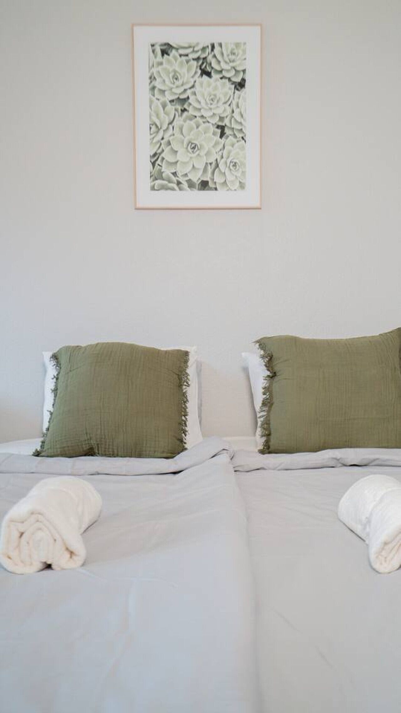 Un letto matrimoniale con biancheria bianca, due cuscini verdi, un quadro floreale incorniciato sulla parete e un asciugamano bianco arrotolato sul letto.