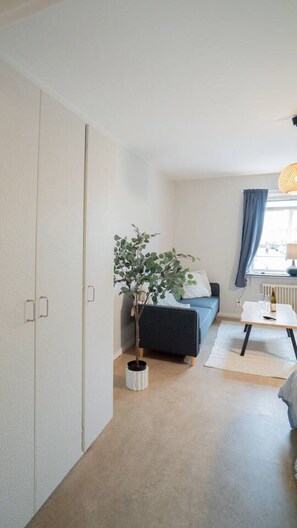 Una habitación con cama, una planta, una mesa con una botella y una ventana con cortinas.