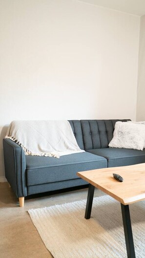 Una sala de estar con un sofá azul oscuro, una manta blanca, una mesa de centro de madera y un control remoto encima.