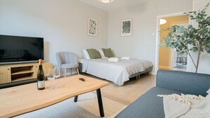 A modern living room with a sofa, a bed, a TV, a wooden table with a bottle and glasses, and a plant.
