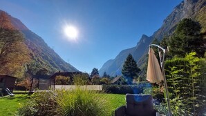Un paysage montagneux sous un ciel dégagé, une maison avec une cheminée bien visible et un jardin avec une chaise et un parasol.