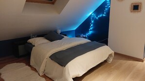 Une chambre avec un plafond en pente, un lit recouvert d’une couette blanche et grise, un plancher en bois, un tapis blanc et des guirlandes lumineuses accrochées au mur.