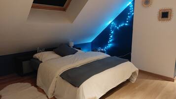 A bedroom with a sloped ceiling, a bed with a white and gray duvet, a wooden floor, a white rug, and string lights on the wall.