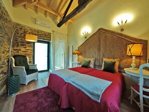 Ein Schlafzimmer mit Steingemäuer, einem Bett mit roter Bettwäsche, einem Sessel, einer Lampe und einer Decke mit freiliegenden Balken.