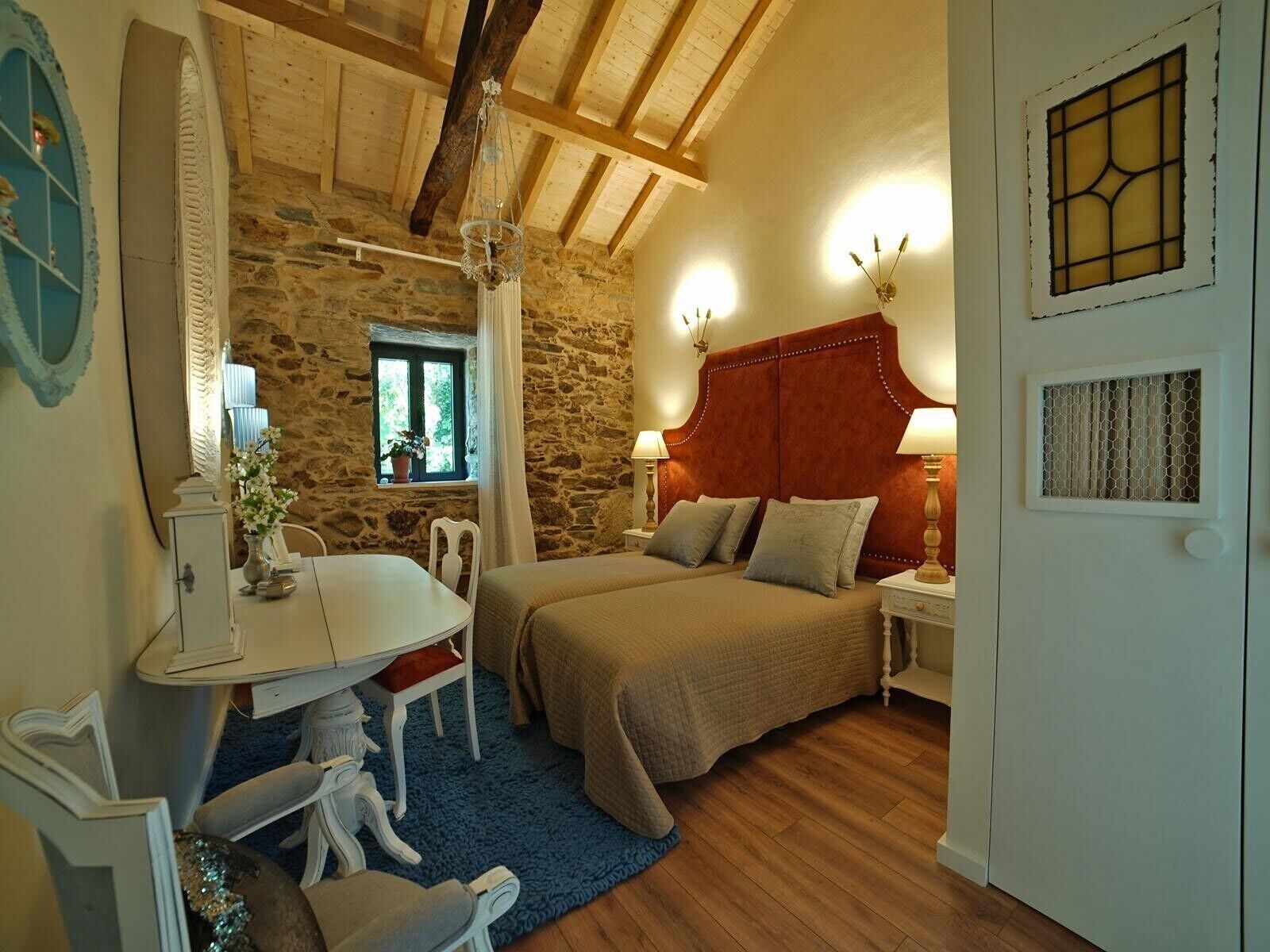 Een slaapkamer met een stenen muur, houten balken in het plafond, een bed met een rode hoofdbord, een nachtkastje met een lamp, en een raam met uitzicht op groen.