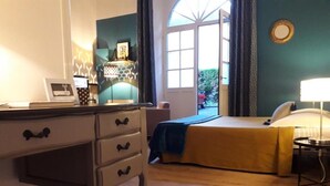 Een slaapkamer met een bed, een bureau met lades, een stoel, een lamp en een raam met gordijnen.