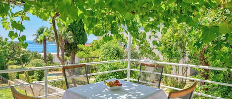 A table with a white tablecloth and a bowl of fruit, surrounded by chairs, with a view of greenery and a body of water in the background.