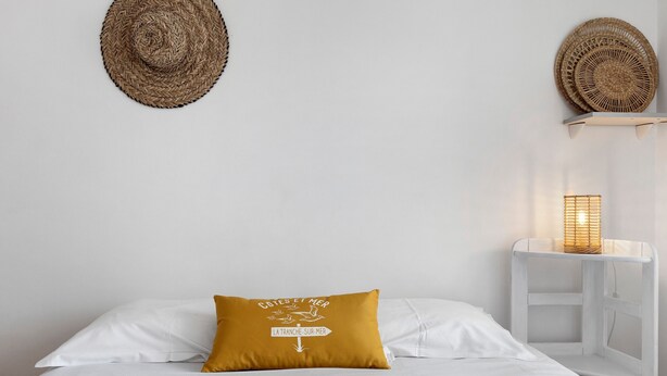 A bed with white bedding, a mustard-colored pillow, a blue towel, and a wall-mounted woven basket. - Victor Studio – 200 m from the Beach (La Tranche-sur-Mer)