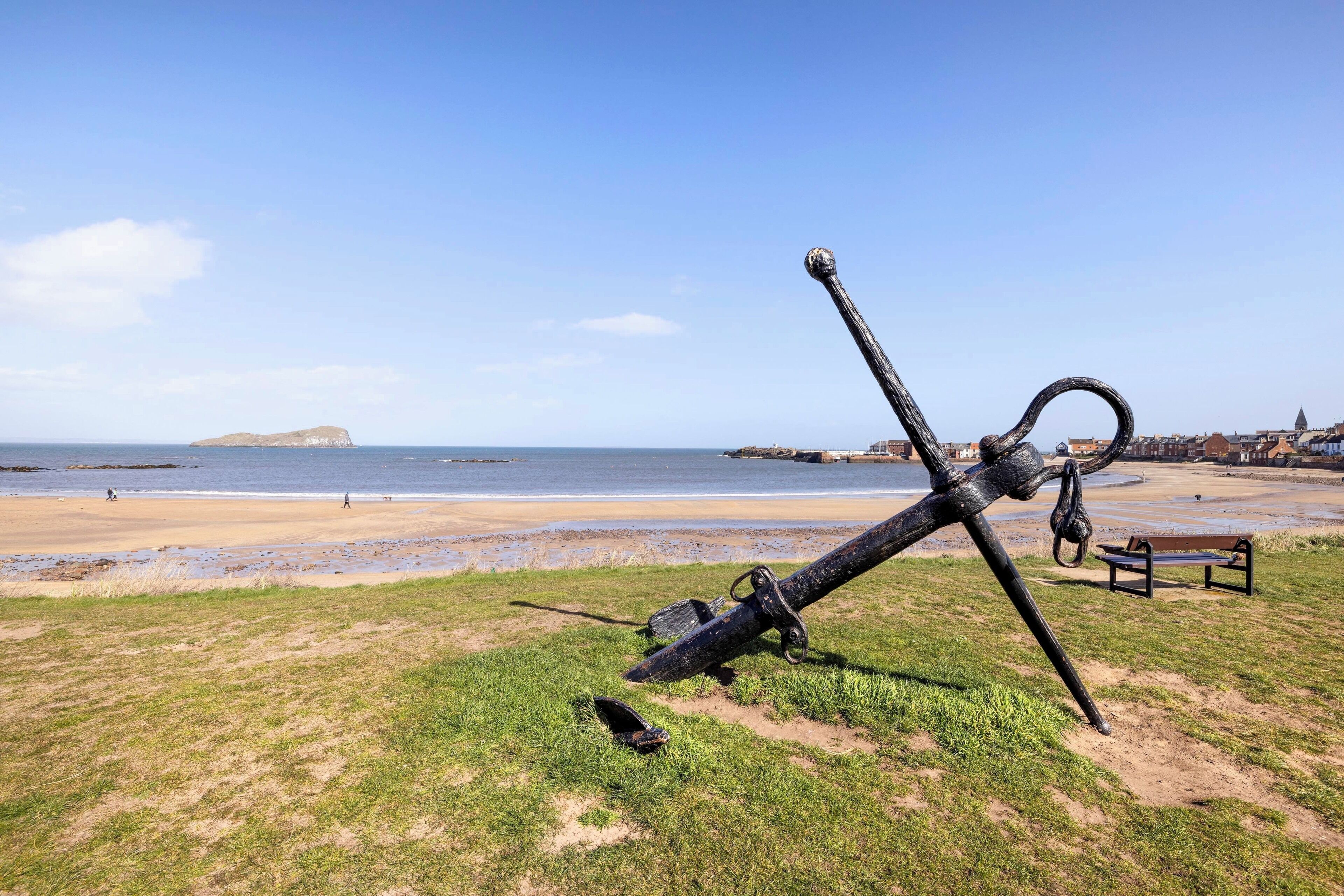 Een strand met een grote ankersculptuur, een bankje en een eiland in de verte.