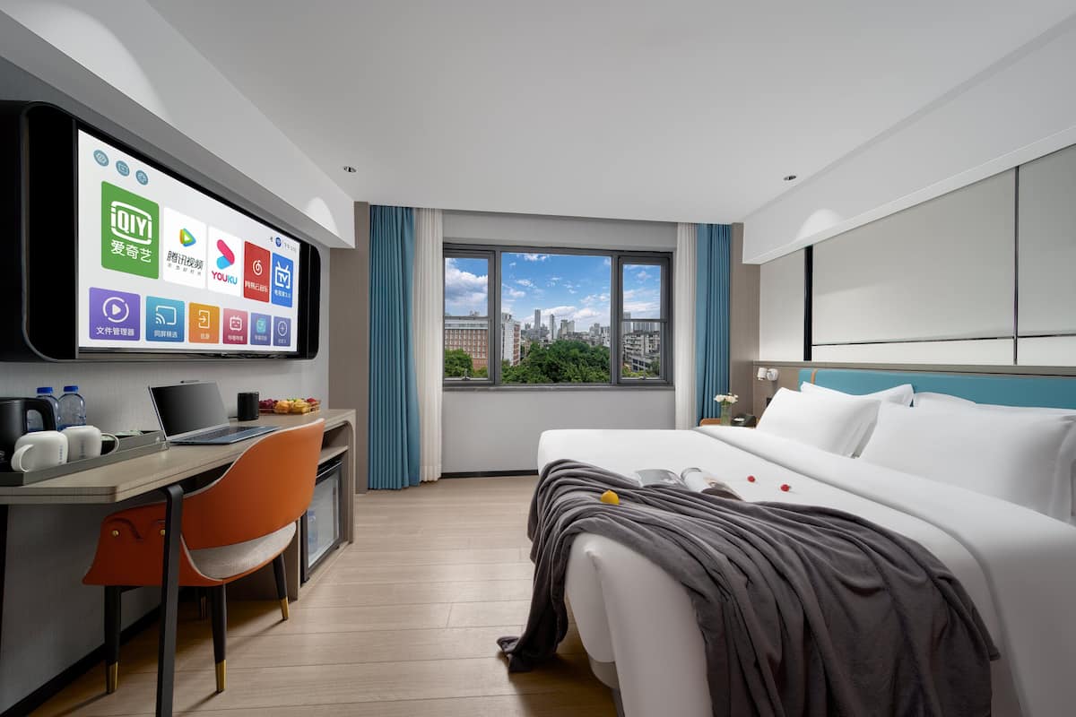 A hotel room with a bed, desk, chair, TV, and a window with a city view.