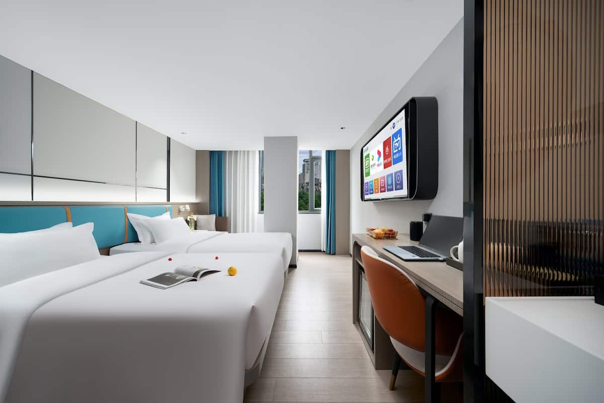 A modern hotel room with two beds, a desk, a television, and a view of the city.