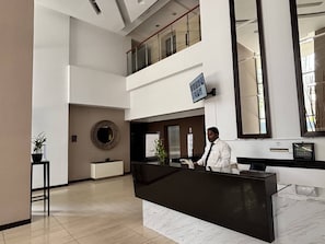 A hotel reception area with a black reception desk, a large mirror, a TV mounted on the wall, and a potted plant.