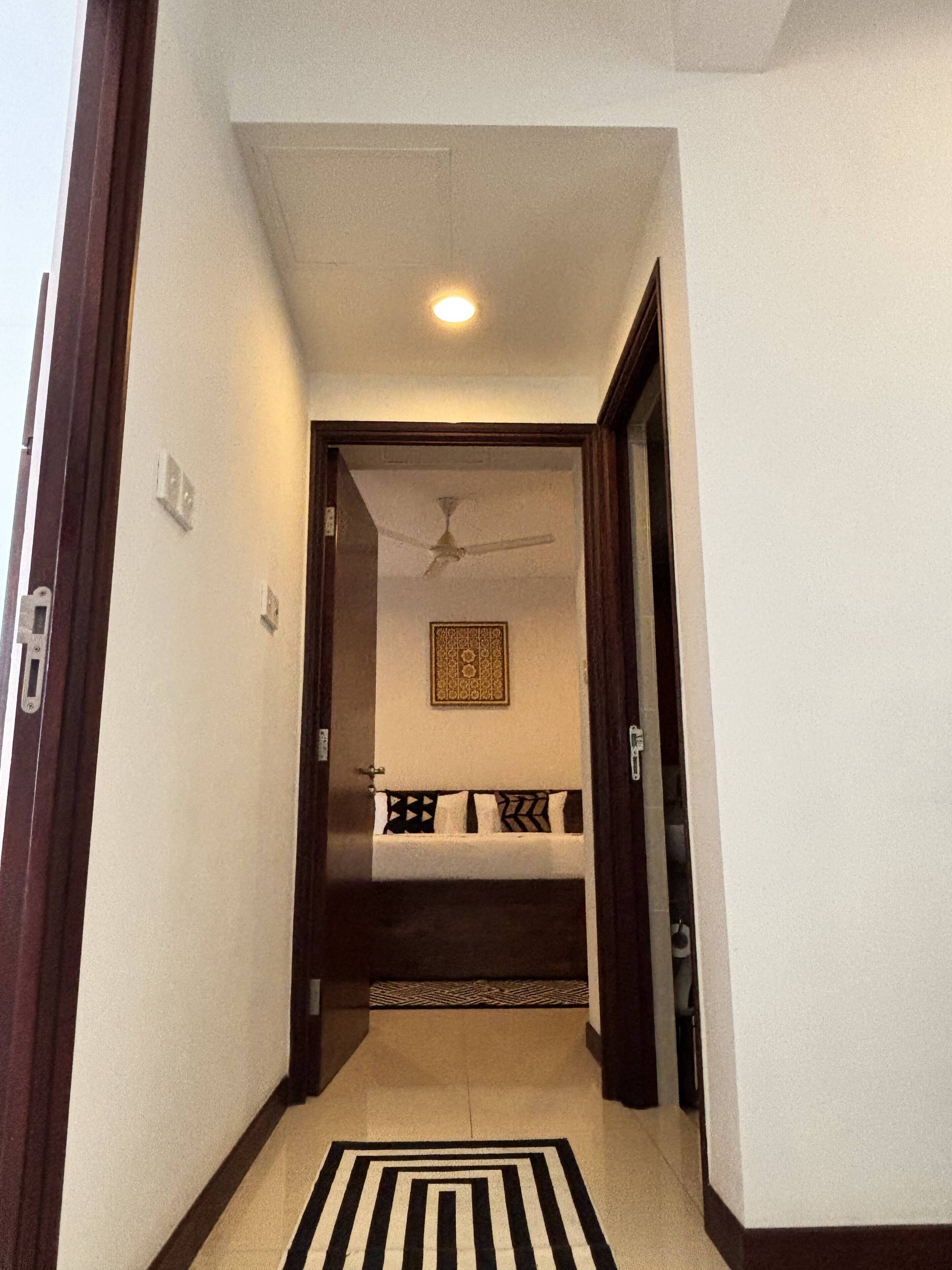A narrow hallway leading to a bedroom with a ceiling fan, a patterned rug, and a bed with patterned pillows.