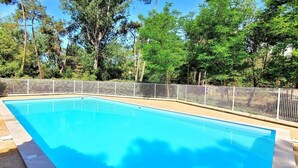 A rectangular swimming pool with a tiled edge, surrounded by a white metal fence, set against a backdrop of tall trees and clear sky. - Forest Apartment – Shared Pool (La Tranche-sur-Mer)