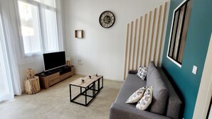 A living room with a sofa, TV, coffee table, and a wall-mounted clock. - Forest Apartment – Shared Pool (La Tranche-sur-Mer)