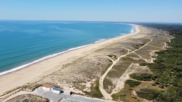 An aerial view of a coastal area with a beach, a road, and a building with a fenced parking area. - Forest Apartment – Shared Pool (La Tranche-sur-Mer)