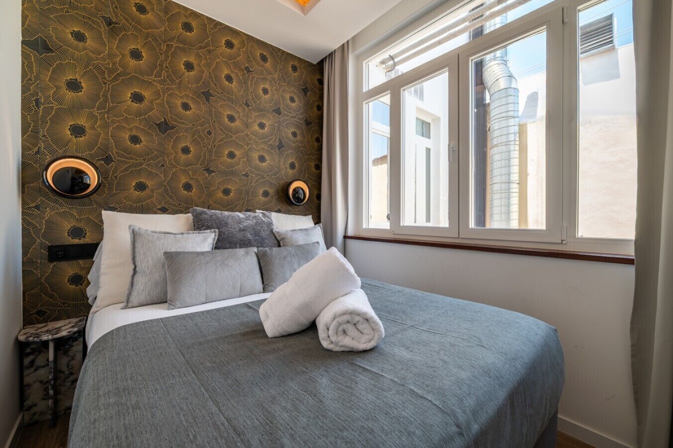 A neatly made bed with a grey blanket and pillows, a round bedside table, a patterned wallpaper, and a window with a view of buildings.