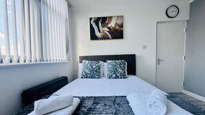A bedroom with a bed, pillows, a painting, a clock, and a window with blinds. - Home Studio  Free Parking Prime Location (Slough)