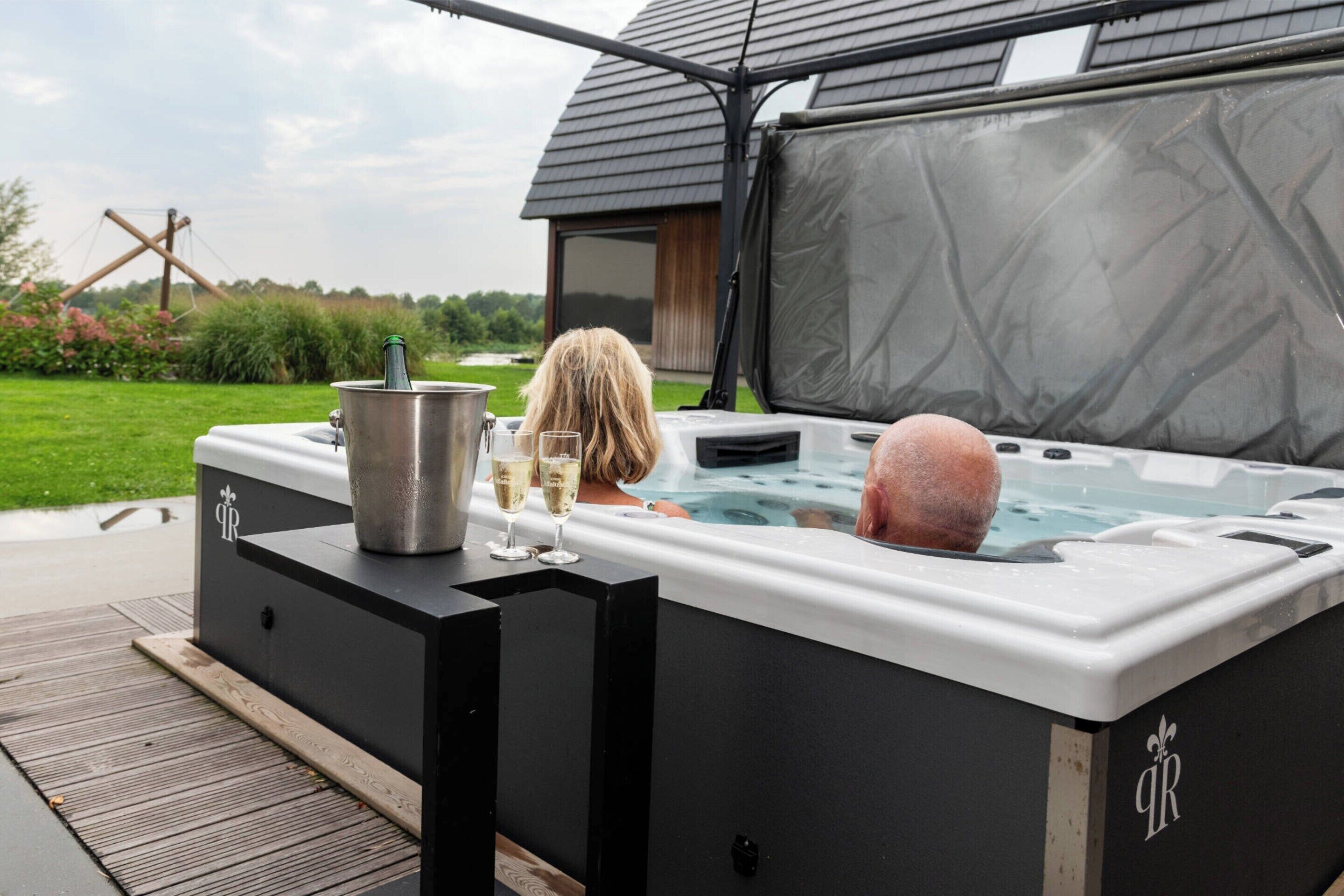 A hot tub with a person sitting inside, a metal container, and two glasses of champagne on the side.