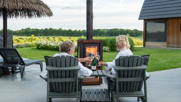 Two people seated at a table with a wood-burning stove, outdoors with a thatched roof and a view of a field. - Private Wellness Haven by the Vecht (Dalfsen)
