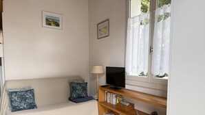 Una sala de estar con sofá, dos almohadas, televisor, lámpara, estantería y ventana con cortinas.